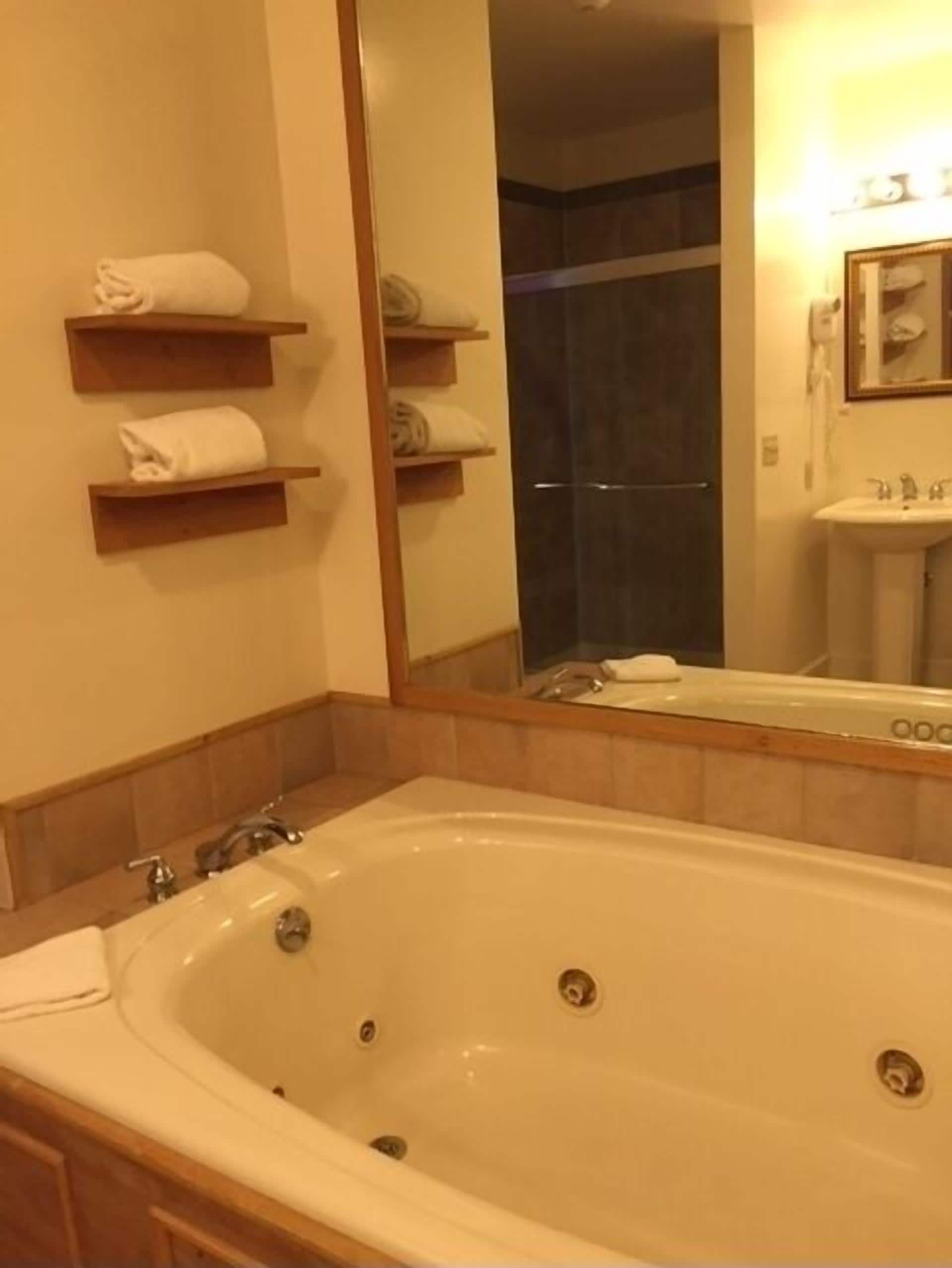 Een badkamer met een groot bad, een wastafel en een spiegel. Handdoeken liggen op planken en er is een douche zichtbaar op de achtergrond.