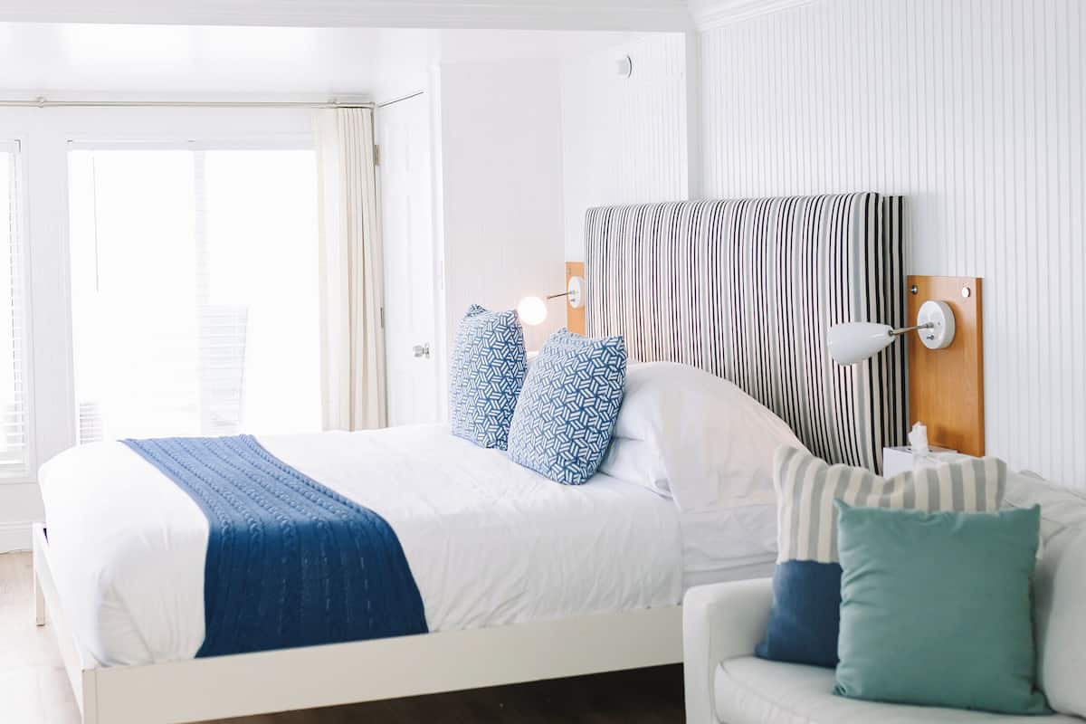 A modern hotel room with a large bed, a sofa, a round ottoman, and a wall-mounted lamp.