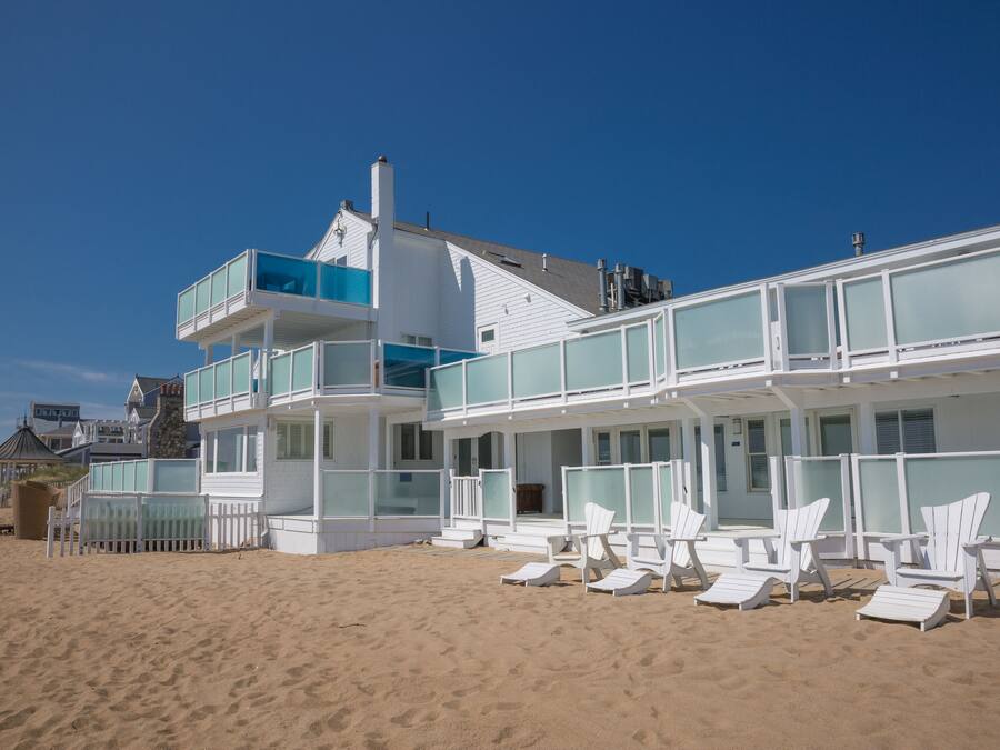 A beachfront property with a multi-story building, a balcony with glass railings, and several beach chairs arranged in front.