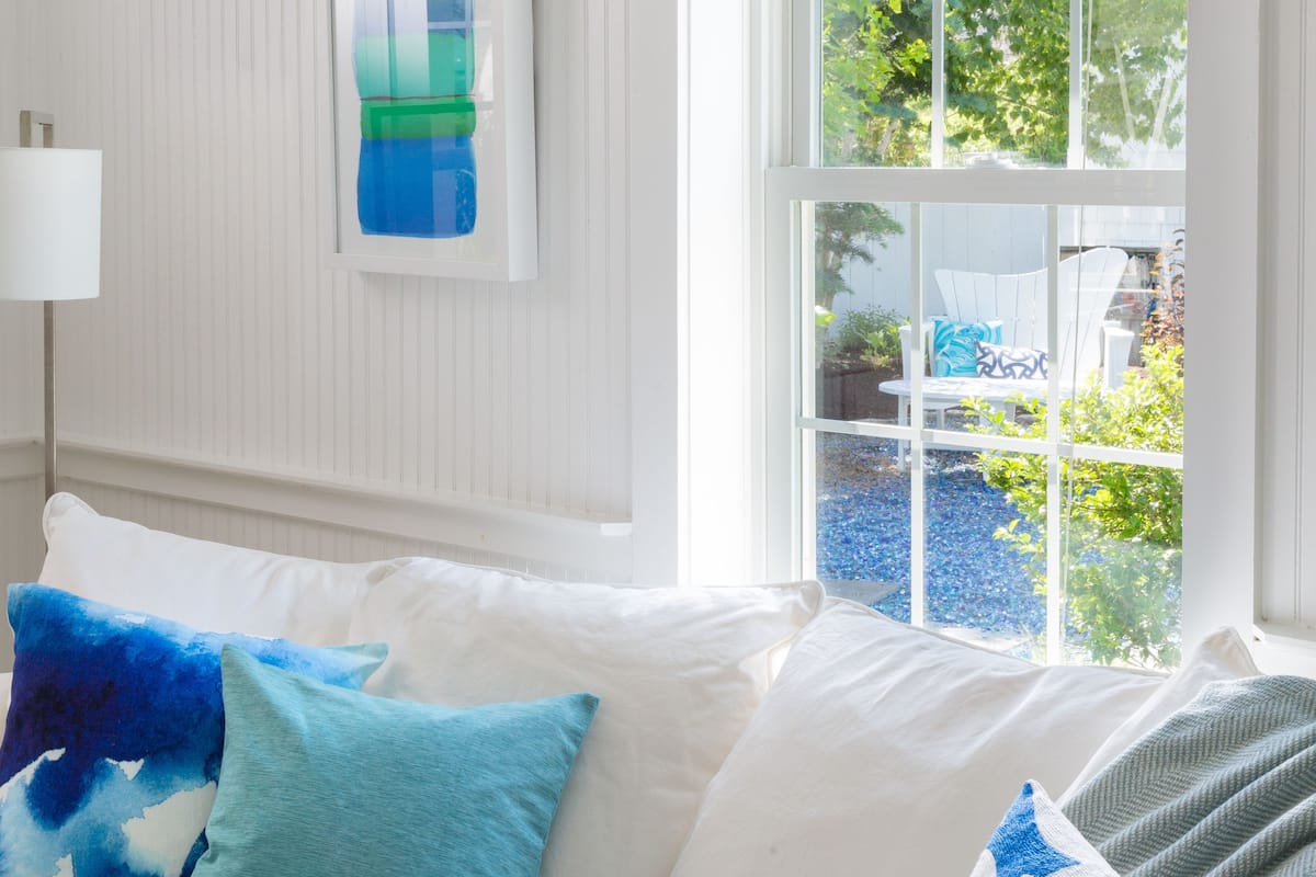 A white sofa with blue and white cushions, a white wall with a framed artwork, and a window with a view of greenery.