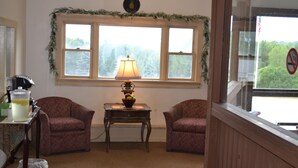 A room with two armchairs, a small table with a lamp, and a window with a view of trees. - Henniker Motel (Henniker)