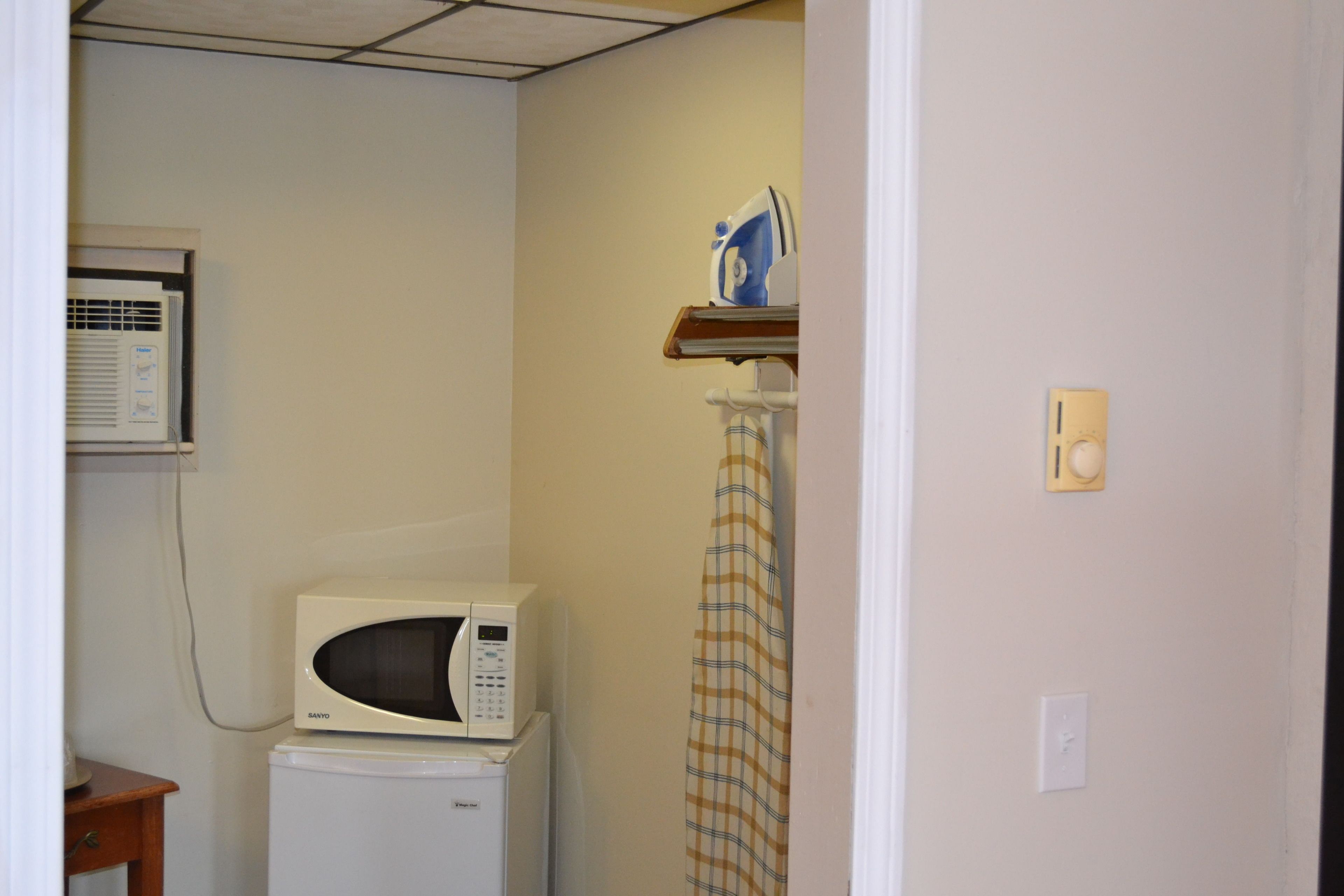 A compact kitchen with a microwave and a small refrigerator, a wall-mounted air conditioner, and a curtain on the door.