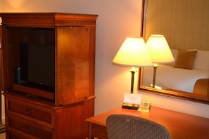 A hotel room with a wooden desk, two lamps, a television, and a mirror reflecting the bed.