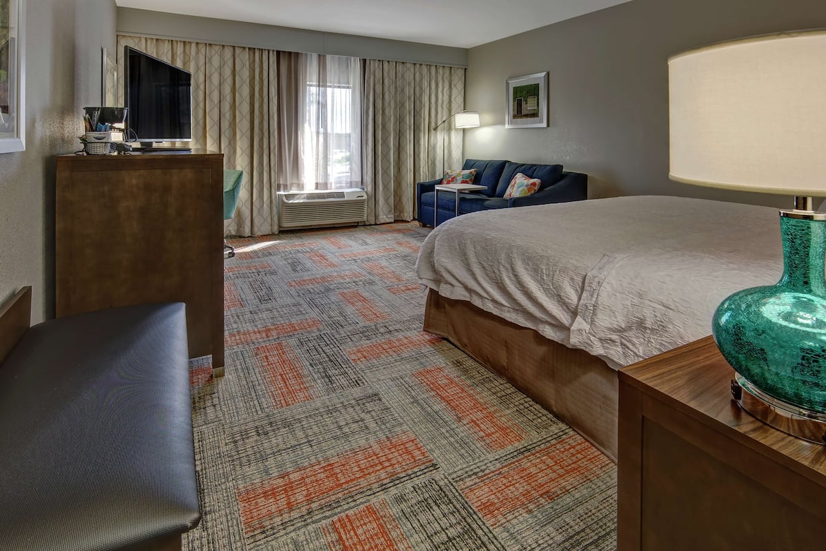 A hotel room with a bed, a desk with a television, a sofa, a lamp, and a patterned carpet.