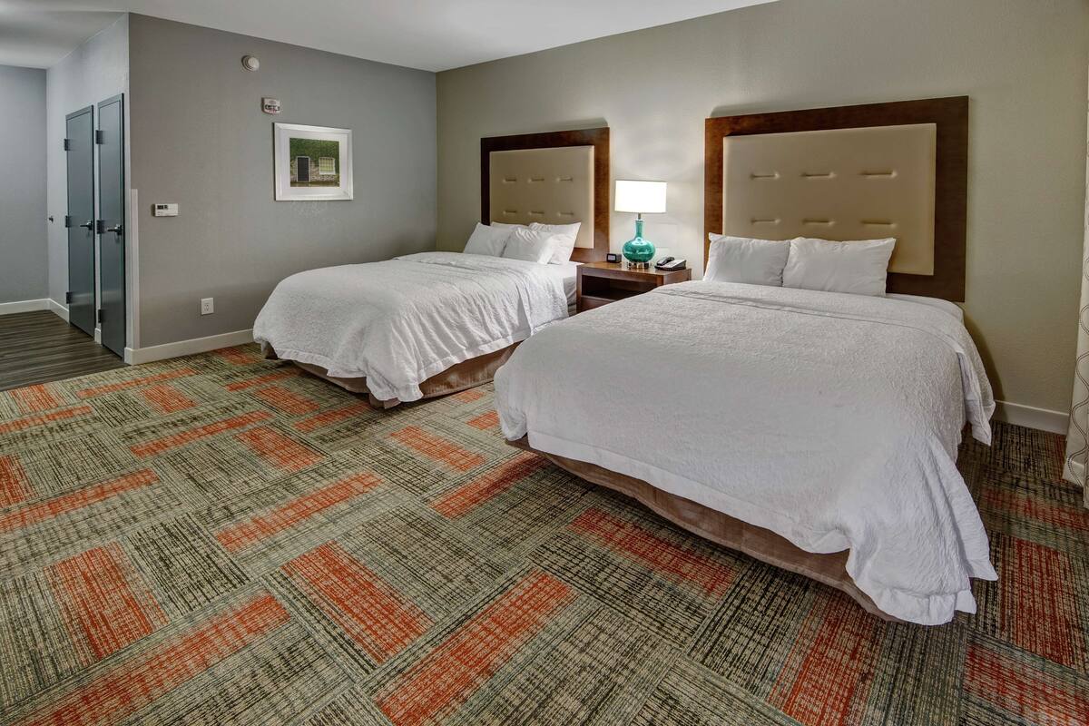A hotel room with two beds, a carpeted floor, and a nightstand with a lamp.