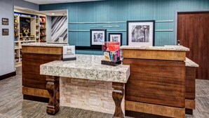 A reception desk with a marble top and wooden base, flanked by shelves stocked with snacks and a 'Welcome to Summerville' sign. - Hampton Inn Summerville SC (Summerville)