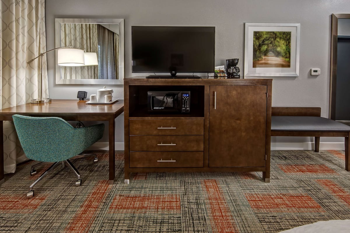 A hotel room with a desk, a chair, a television, a microwave, and a framed picture on the wall.