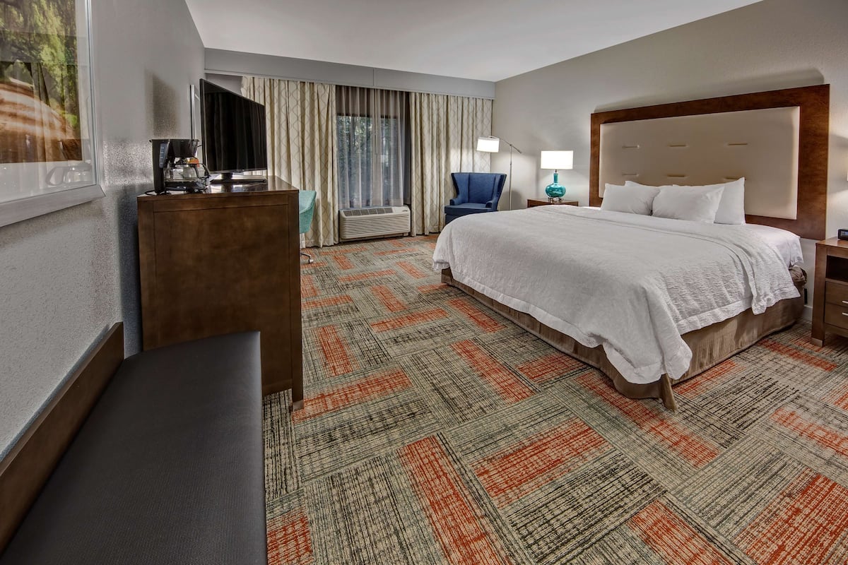 A hotel room with a large bed, a desk with a coffee maker, a chair, a TV, and a window with curtains.