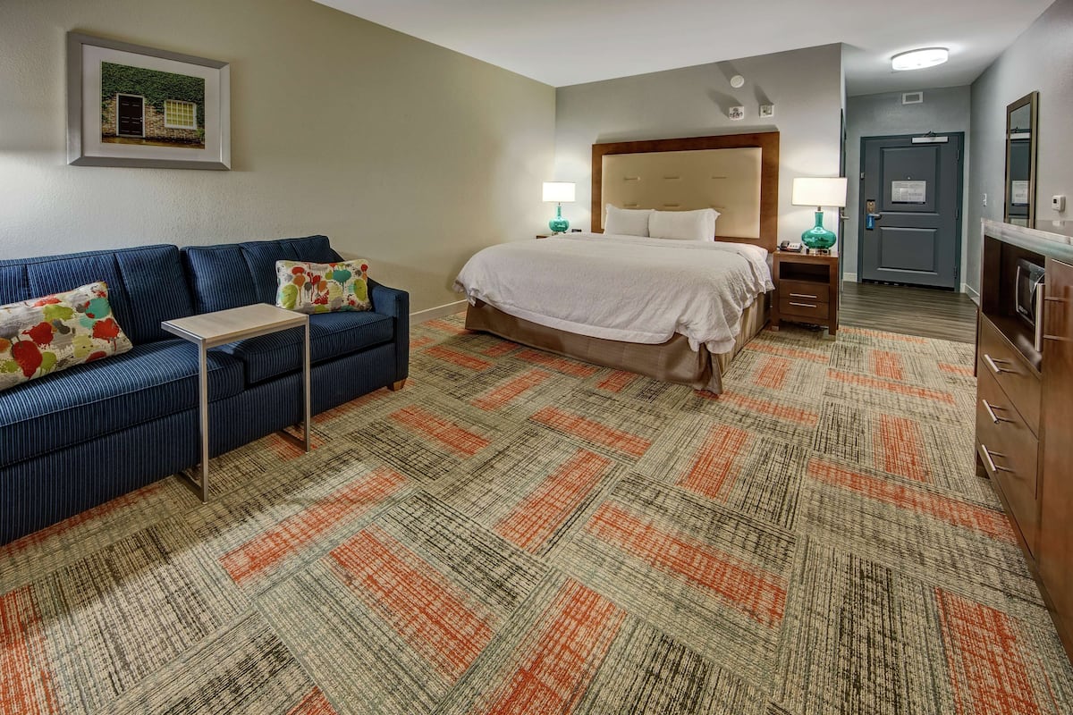 A hotel room with a bed, a sofa, a small table, a lamp, and a kitchenette.