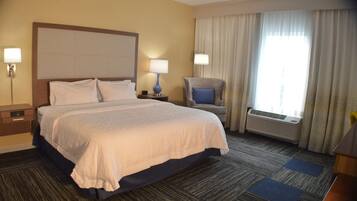 Habitación de hotel con una cama grande, dos mesitas de noche con lámparas, una silla, un aire acondicionado y una ventana con cortinas.