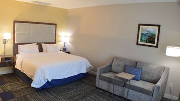 A hotel room with a bed, a gray sofa, a nightstand, a lamp, and a painting on the wall.