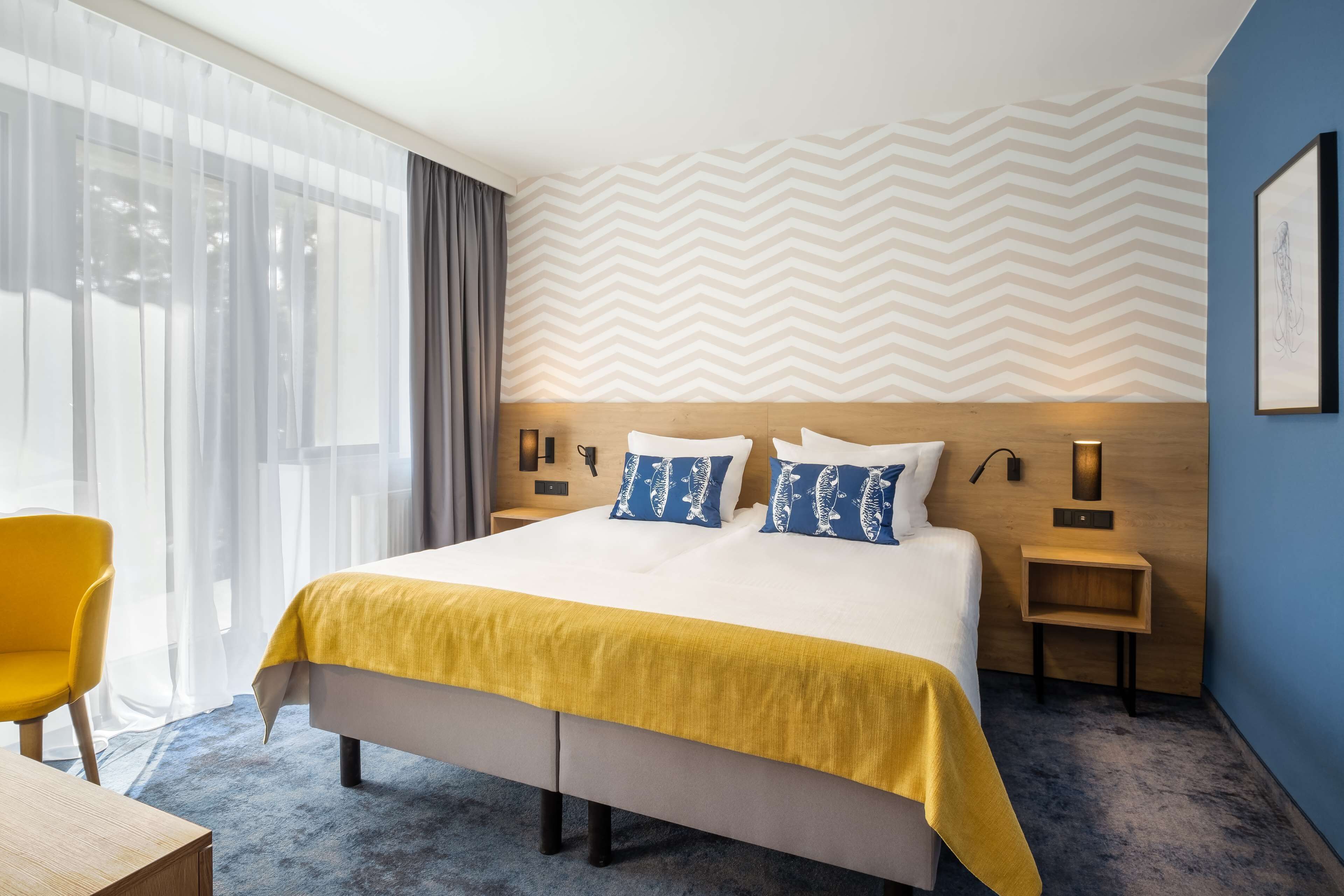 A hotel room with a large bed, a yellow throw blanket, two pillows with a fish pattern, a wooden headboard, bedside tables with lamps, a chair, and a view of the outdoors through sheer curtains.