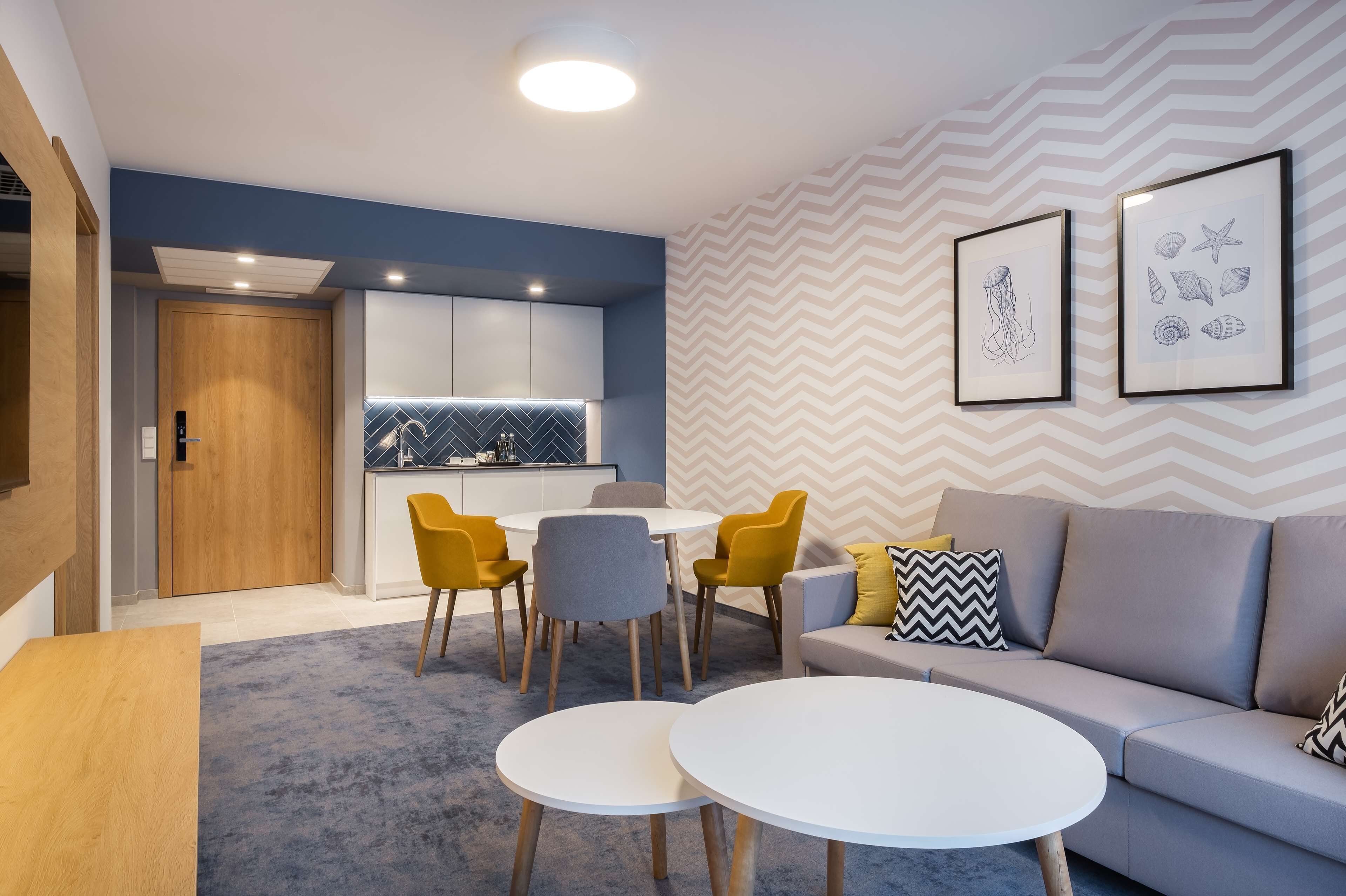 Ein modernes Wohnzimmer mit einem grauen Sofa, gelben Sesseln und einem weißen Couchtisch. Es gibt eine Küchenzeile mit einer blauen Spritzschutzwand und einer Holztür.