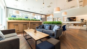 Een moderne woonkamer met een bank, salontafel en barstoelen. Er is een keukenhoek met een televisie en een groot raam.