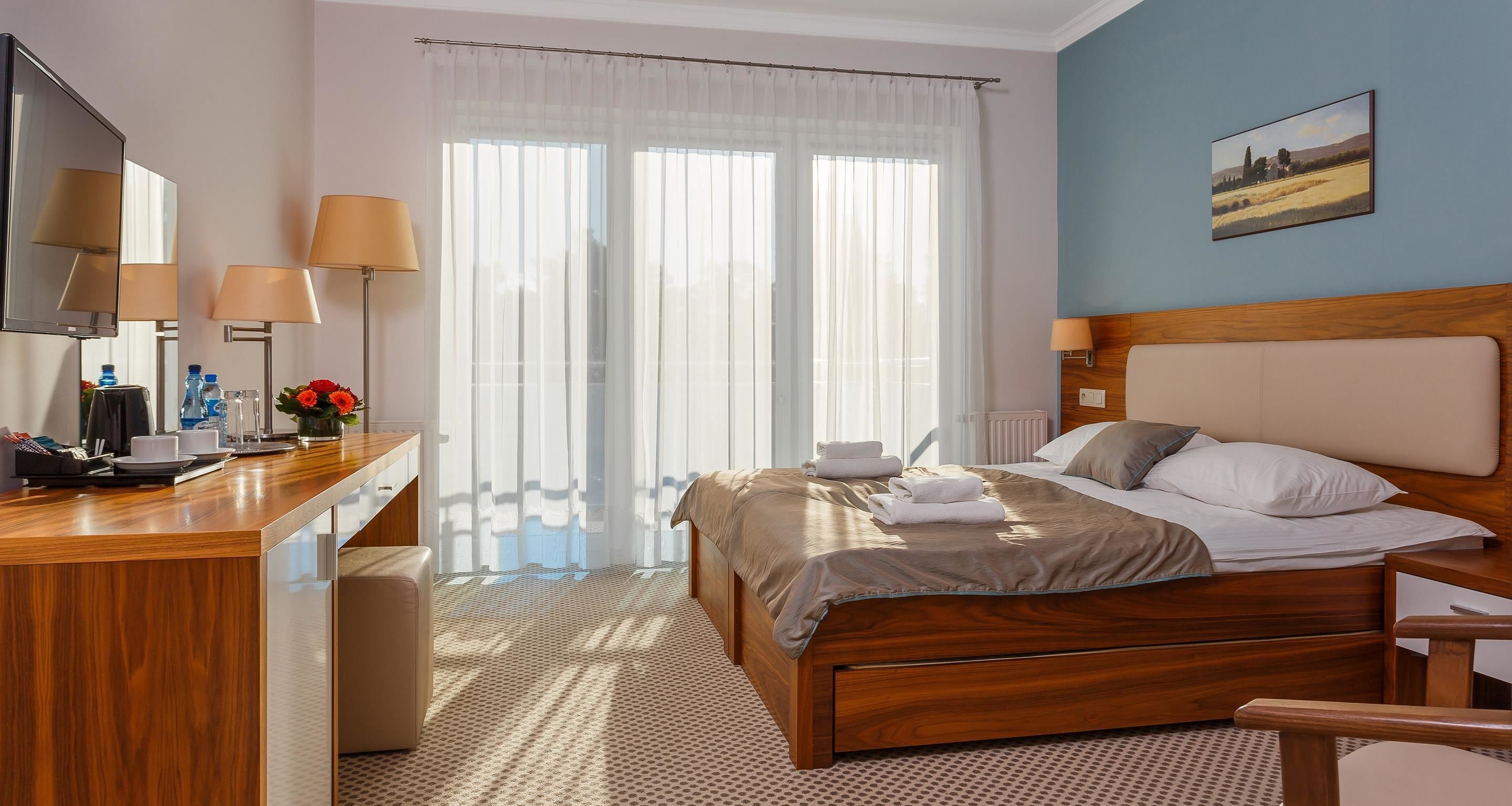 Habitación de hotel con cama de madera, un escritorio con teléfono y lámparas, una ventana con cortinas translúcidas y un cuadro en la pared.