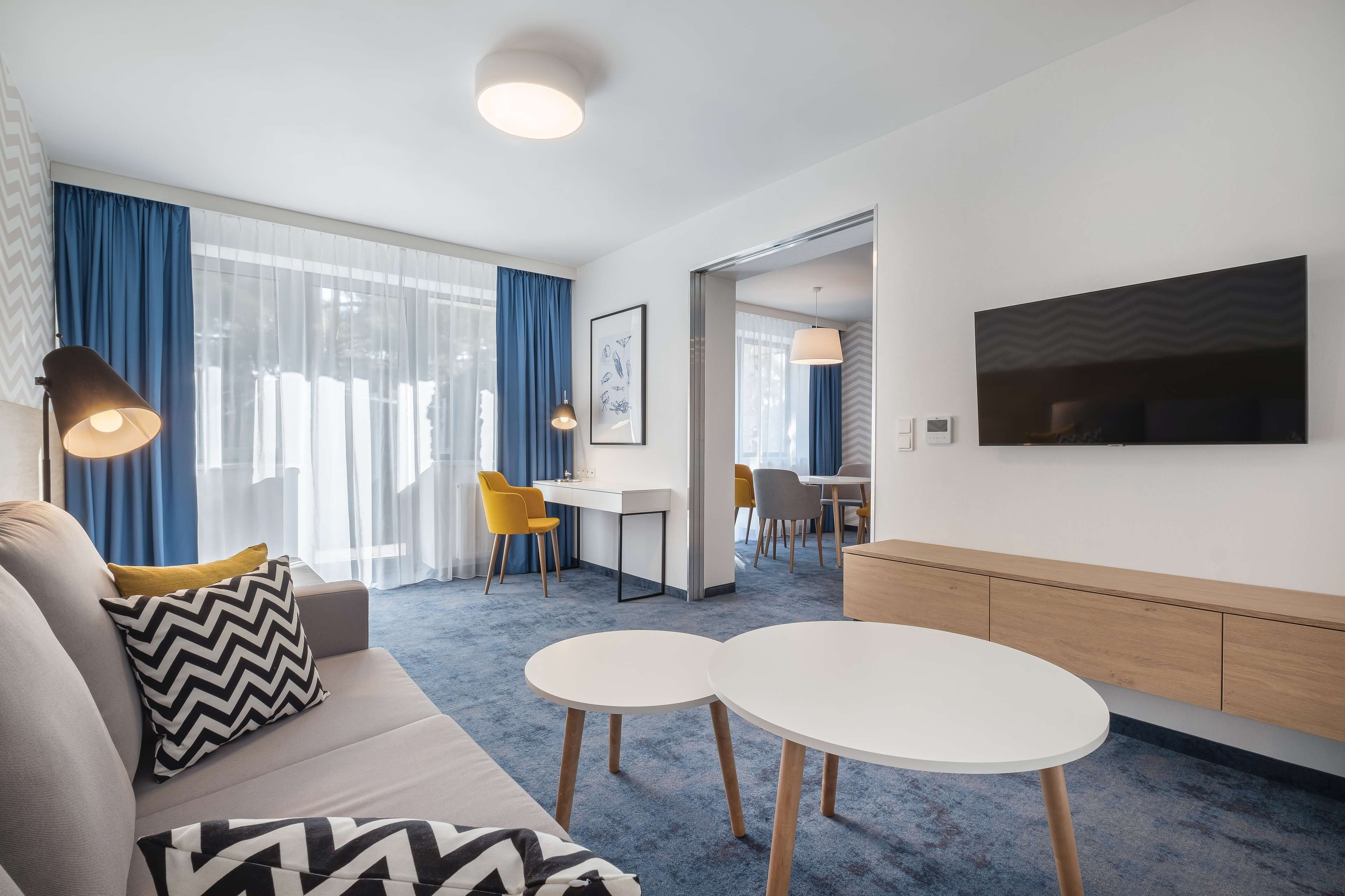 Una habitación de hotel moderna con un sofá, dos mesas redondas, un televisor y un escritorio con sillas.