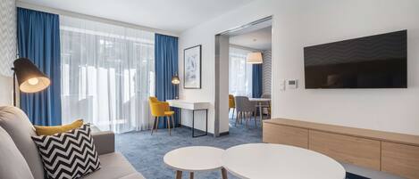 Una habitación de hotel moderna con un sofá, dos mesas redondas, un televisor y un escritorio con sillas.