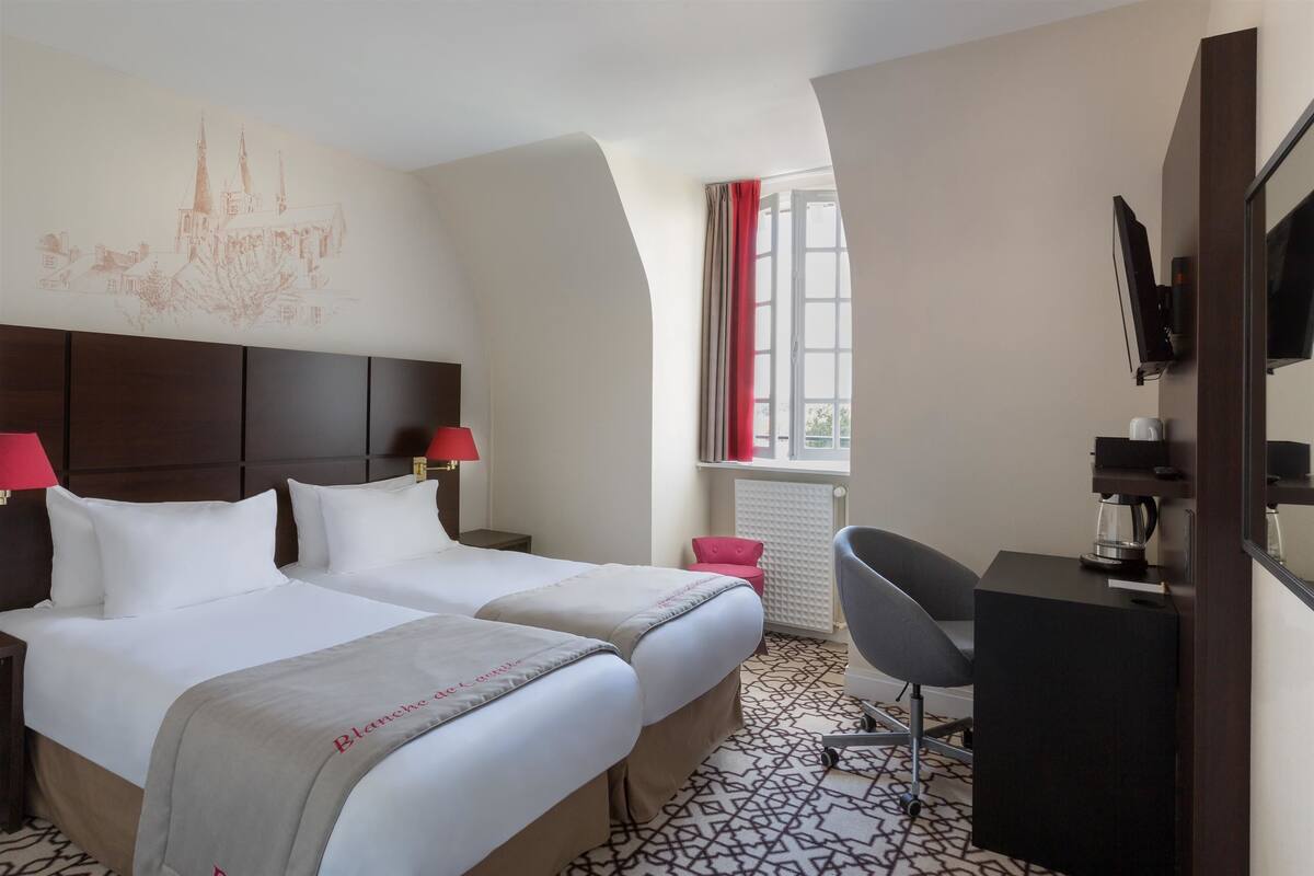 Quarto de hotel com duas camas, uma escrivaninha com cadeira, televisão e janela com cortinas.