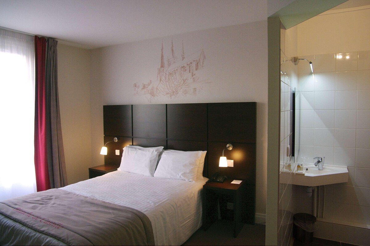 Habitación de hotel con cama, dos lámparas de noche, mesita de noche con teléfono y baño con espejo y lavamanos.