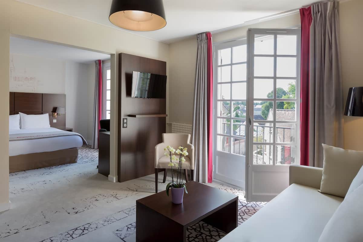 Quarto de hotel com cama, sofá, mesa de centro com planta, televisão e varanda com cortinas.