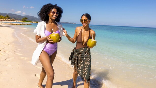 Dos personas caminando sobre la playa sosteniendo cocos.