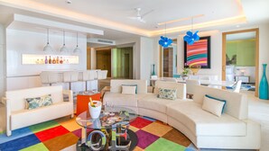 A modern living room with a white sofa, a glass coffee table, and a colorful rug. There are pendant lights, a ceiling fan, and a large abstract painting on the wall.