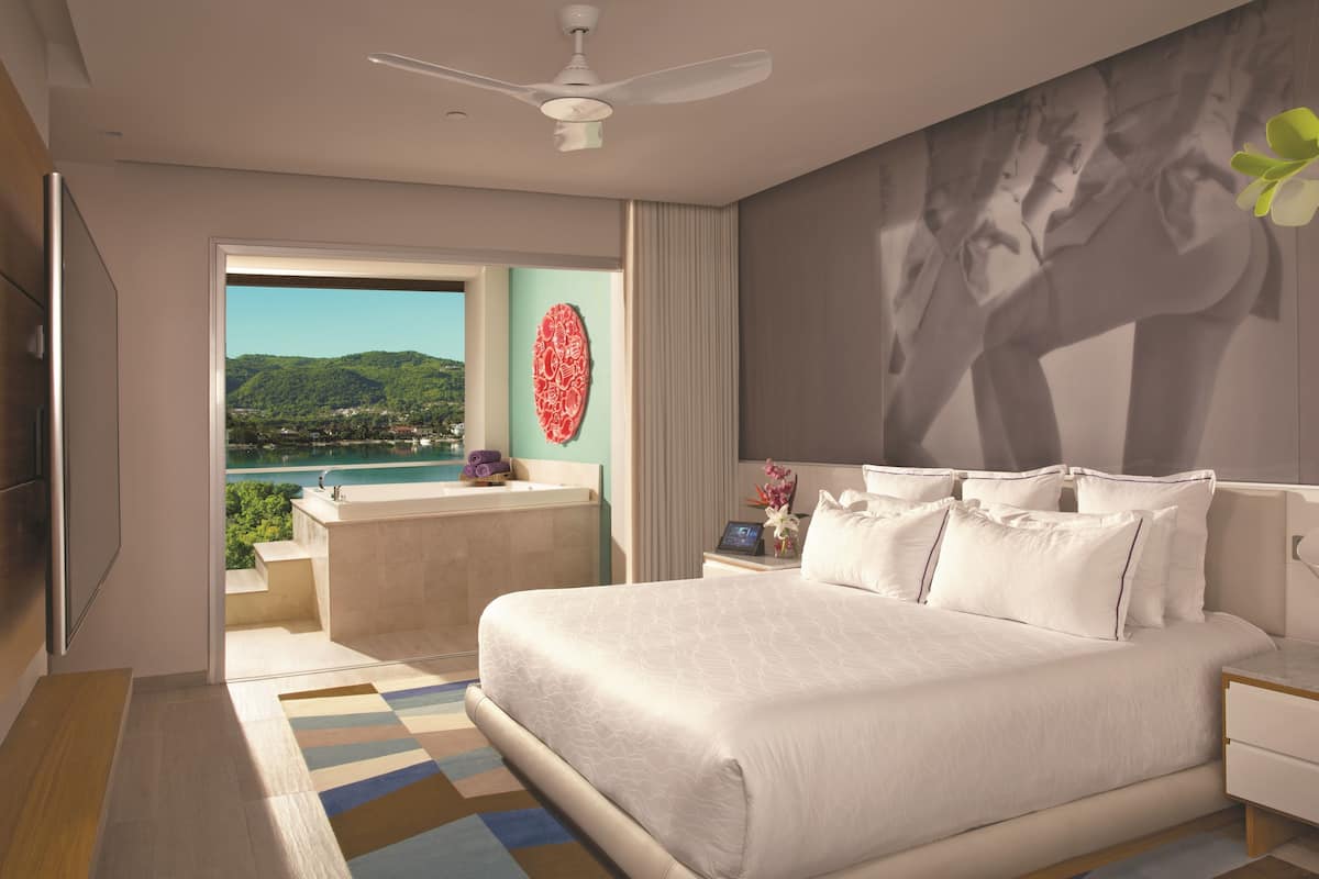 Een moderne hotelkamer met een groot bed, een badkamer met bad en een uitzicht op een meer, plus een plafondventilator.