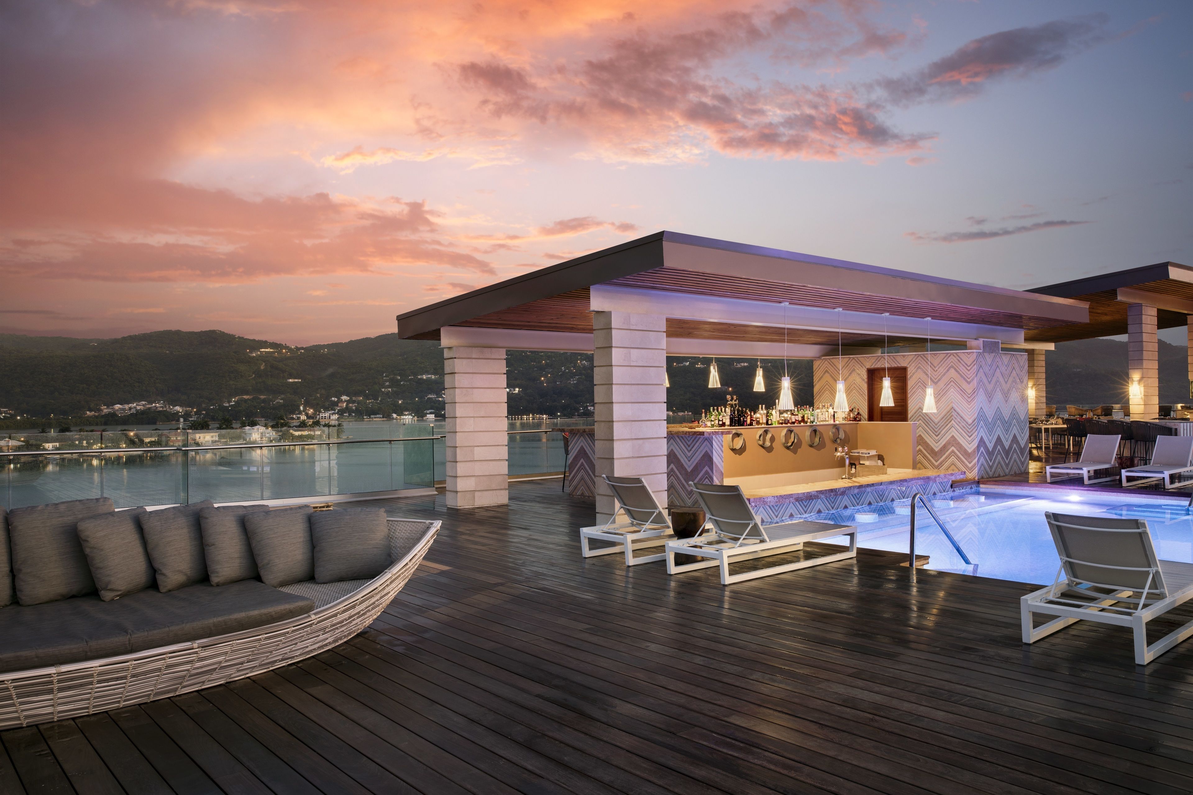Un bar en la azotea con piscina, sillones de descanso y una zona de bar con vistas a la ciudad al atardecer.