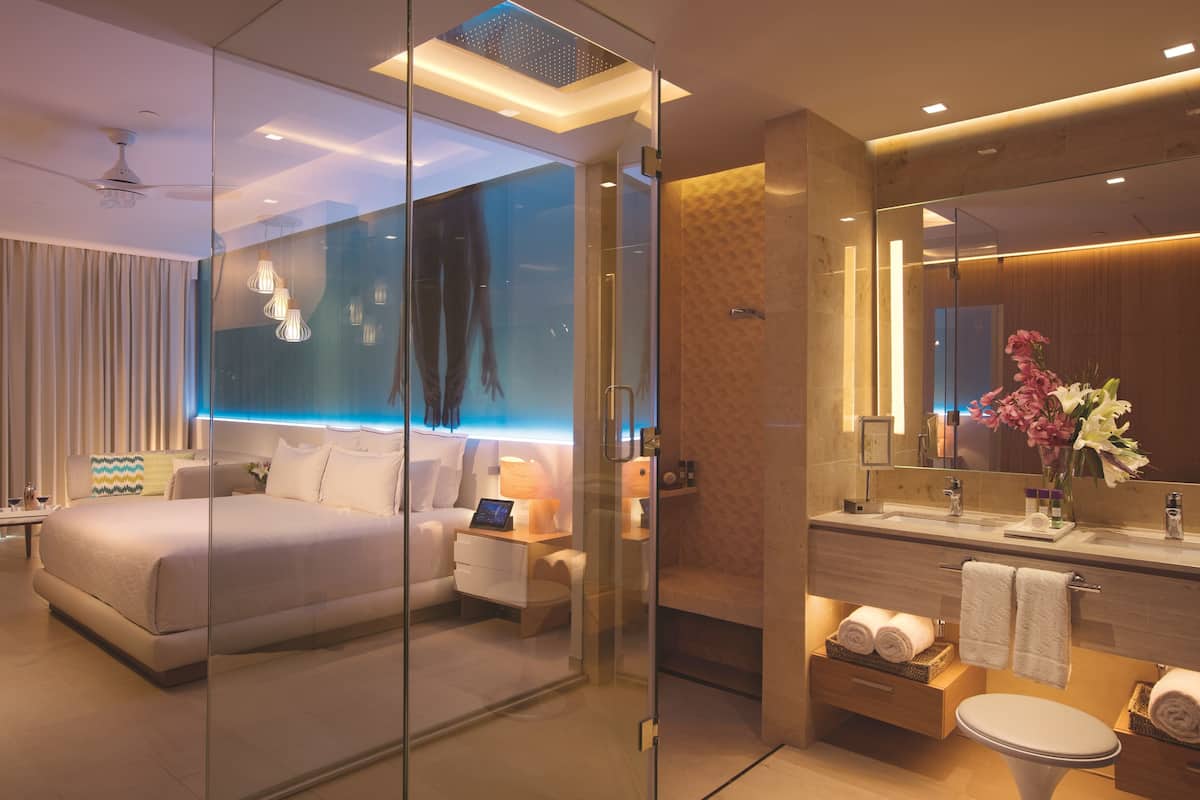 Een moderne hotelkamer met een glazen scheiding tussen de slaapkamer en de badkamer. De slaapkamer heeft een groot bed, een nachtkastje met een lamp en een tv aan de muur. De badkamer is voorzien van een moderne wastafel met spiegel, een toilet en een handdoekhouder.