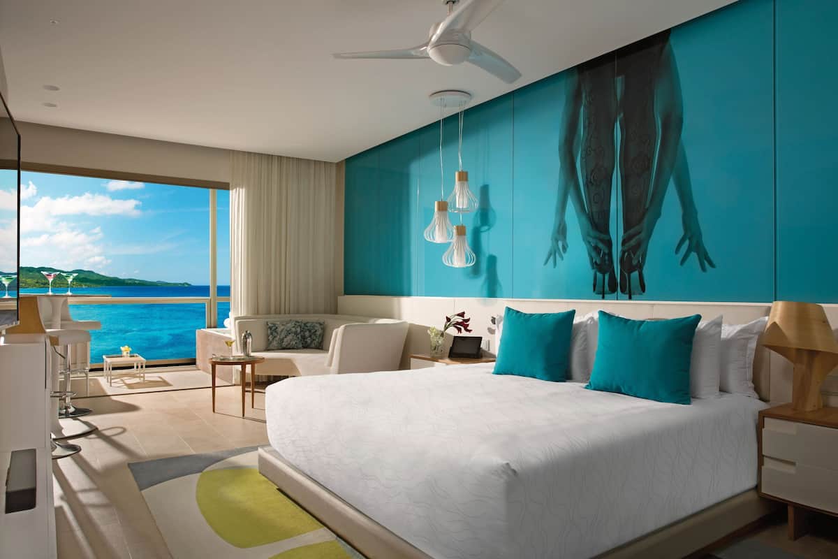 Een moderne hotelkamer met een groot bed, een plafondventilator, uitzicht op zee en een zithoek met uitzicht.