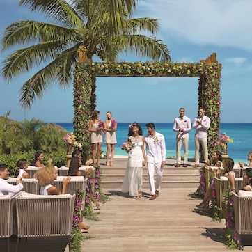 A wedding ceremony on a wooden deck with a floral arch and guests seated on wicker chairs.