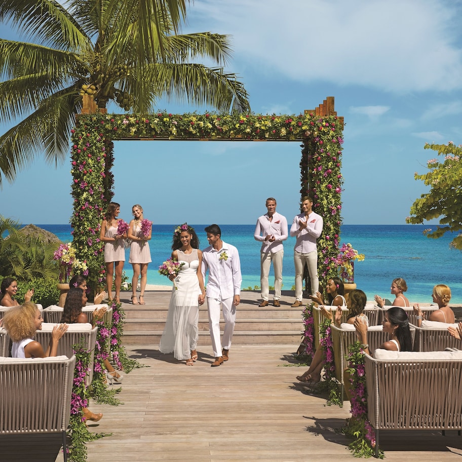 A wedding ceremony on a wooden deck with a floral arch and guests seated on wicker chairs.