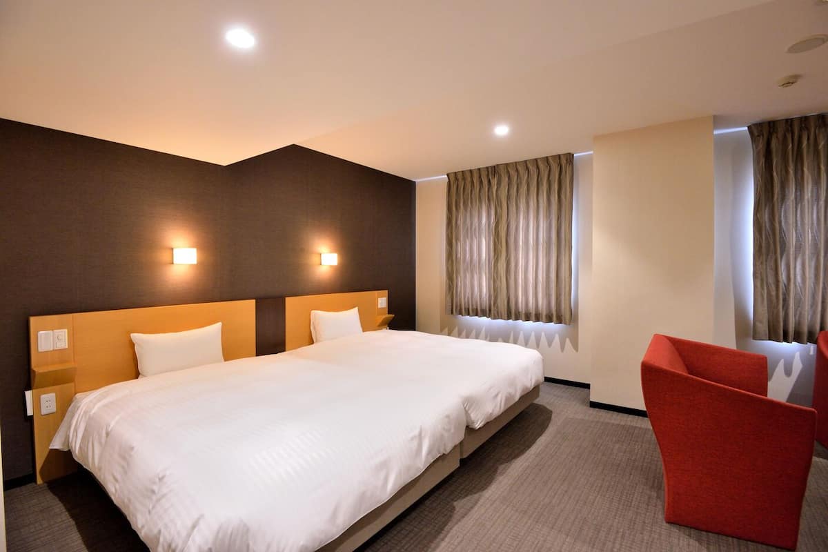 Quarto de hotel com uma cama grande, duas mesas de cabeceira, uma poltrona vermelha e uma janela com cortinas.