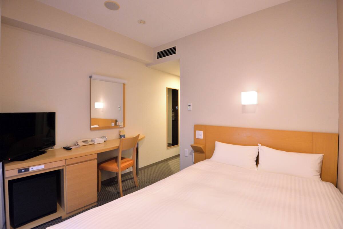 Quarto de hotel com cama, escrivaninha com televisão, uma cadeira e espelho.