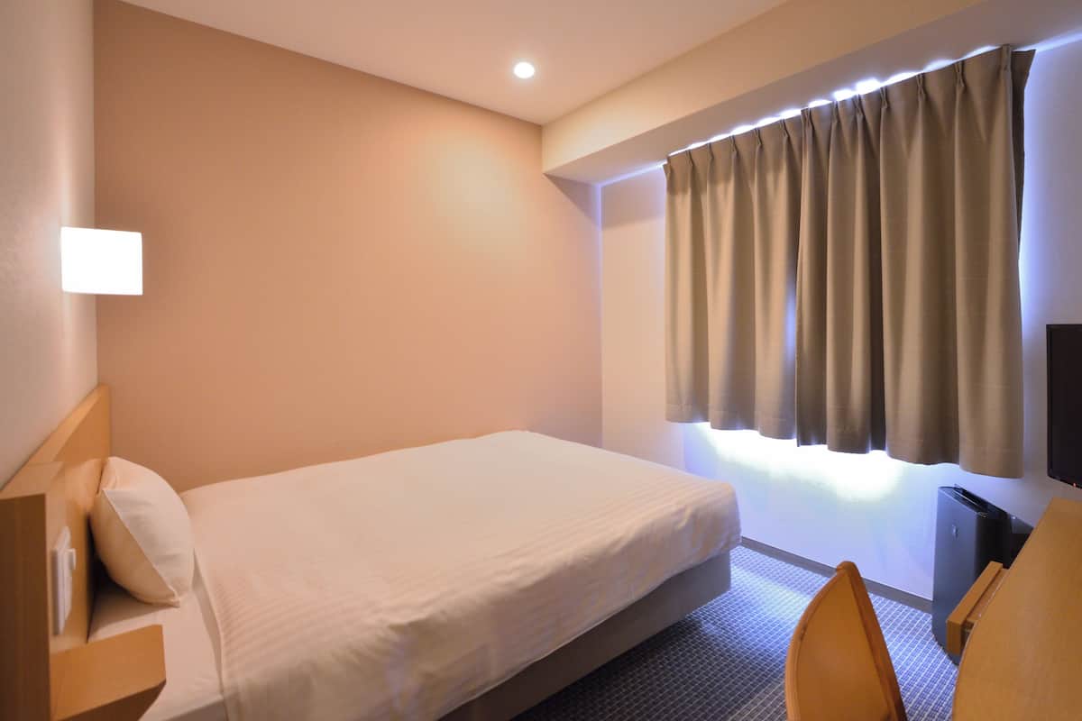 Quarto de hotel com uma cama de solteiro, mesa de cabeceira, cadeira, televisão e janela com cortinas.