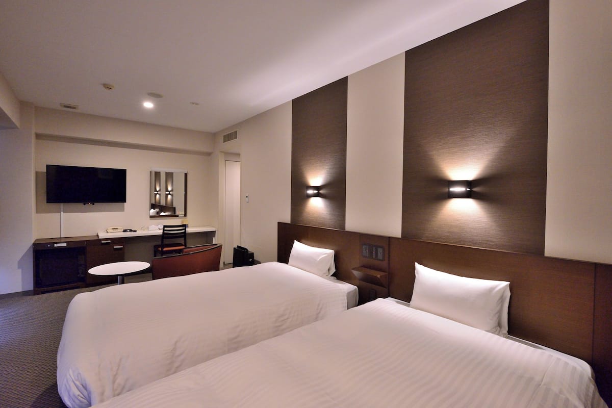 Quarto de hotel com duas camas, uma escrivaninha, uma cadeira, uma televisão e um espelho.