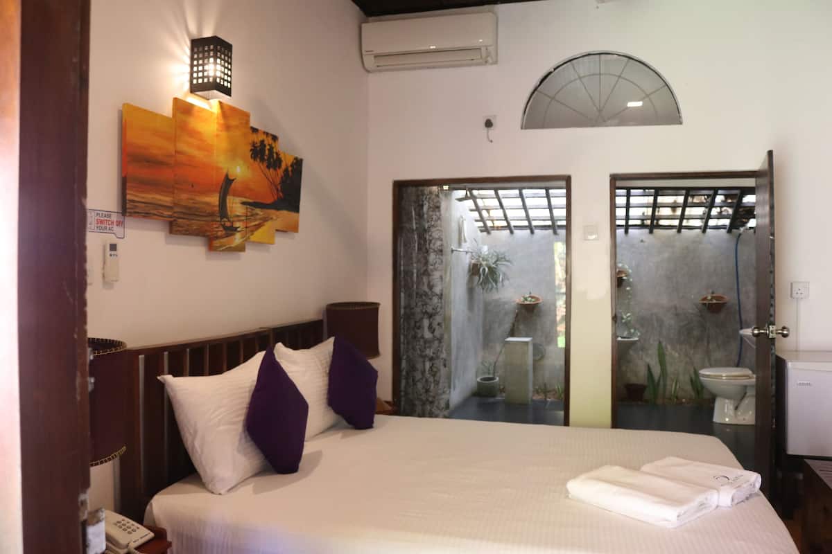 A hotel room with a bed, a nightstand, a lamp, a painting, an air conditioner, a shower area, and a toilet.