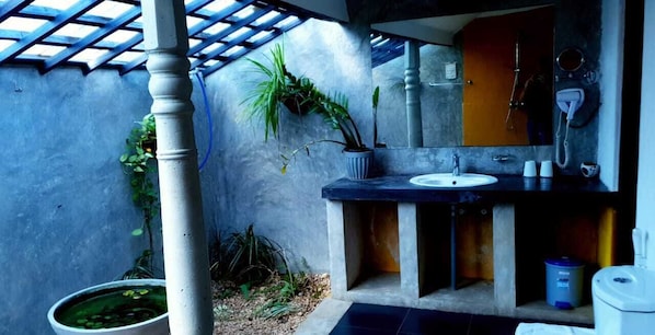 Un baño con lavamanos, espejo y ducha. Hay plantas y una maceta en el suelo.