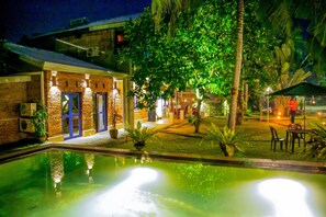 Una vista nocturna de un patio de hotel con piscina, edificios de ladrillo y zonas de estar al aire libre.
