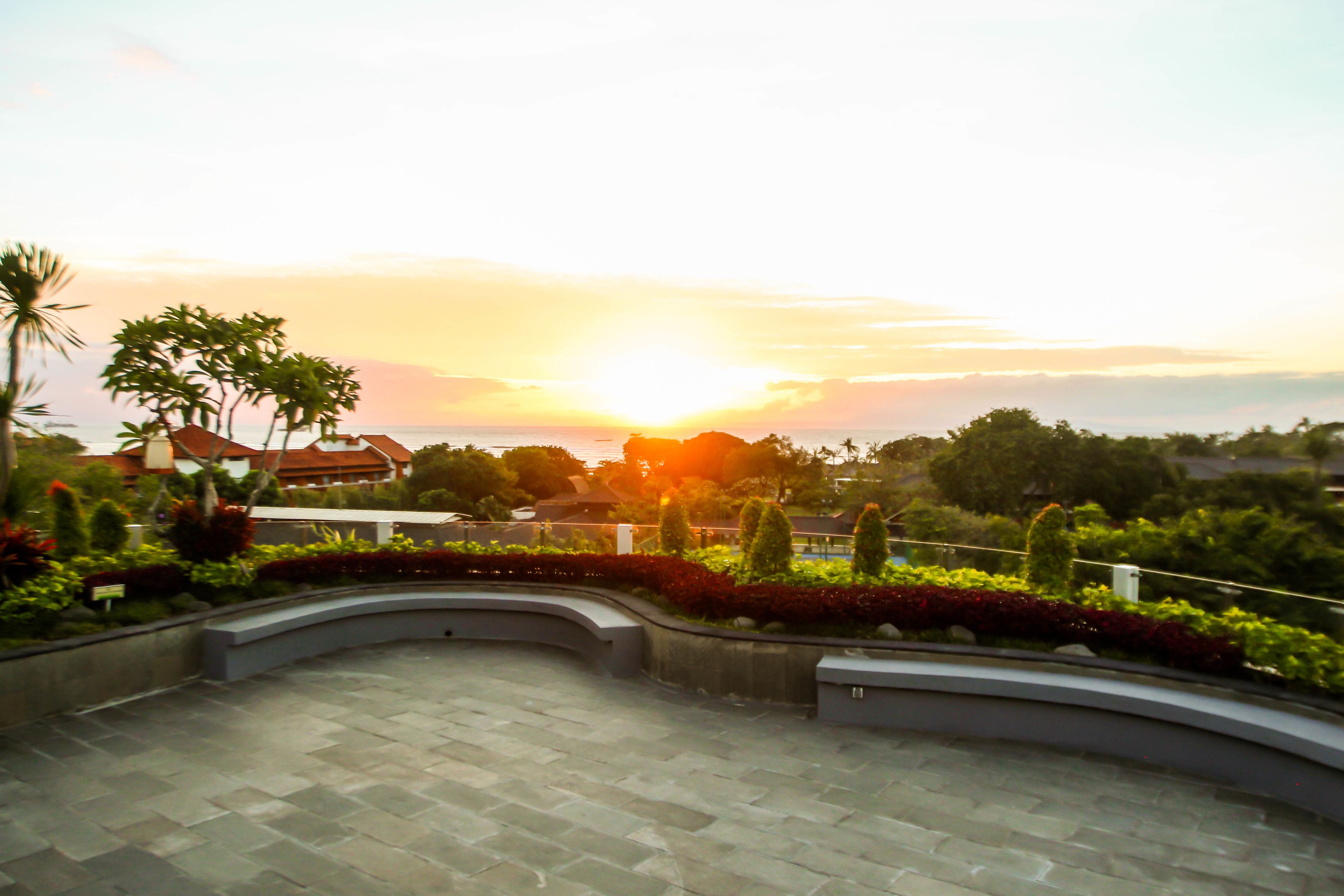 A courtyard with a curved pathway, surrounded by well-maintained greenery and a view of the sunset over a distant landscape.