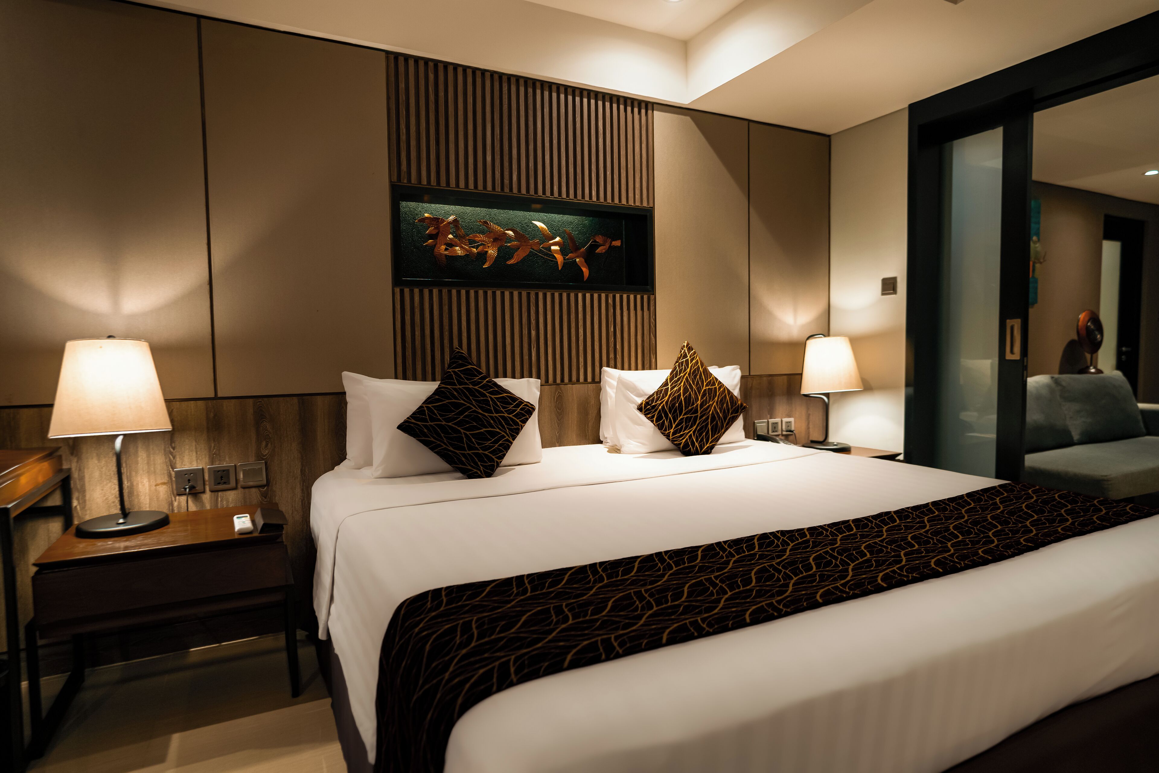 Habitación de hotel con una cama grande, dos mesitas de noche con lámparas, un adorno en la pared y un espejo en la pared derecha.