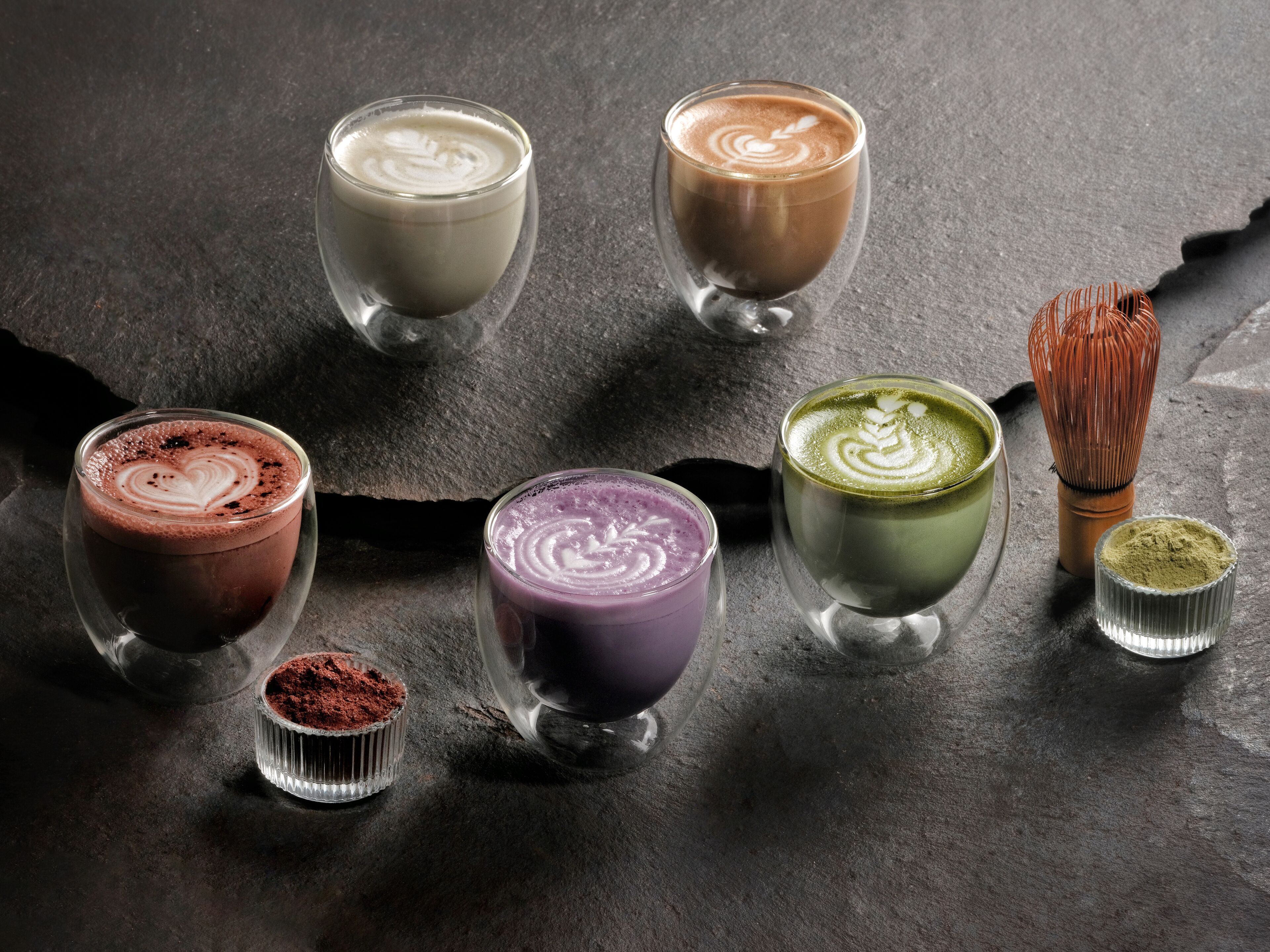 Kuusi eriväristä juomaa tarjoiltuna laseissa, ja jokaisessa on ainutlaatuinen latte art -kuvio, ja ne on asetettu tummanväriselle kivipinnalle.