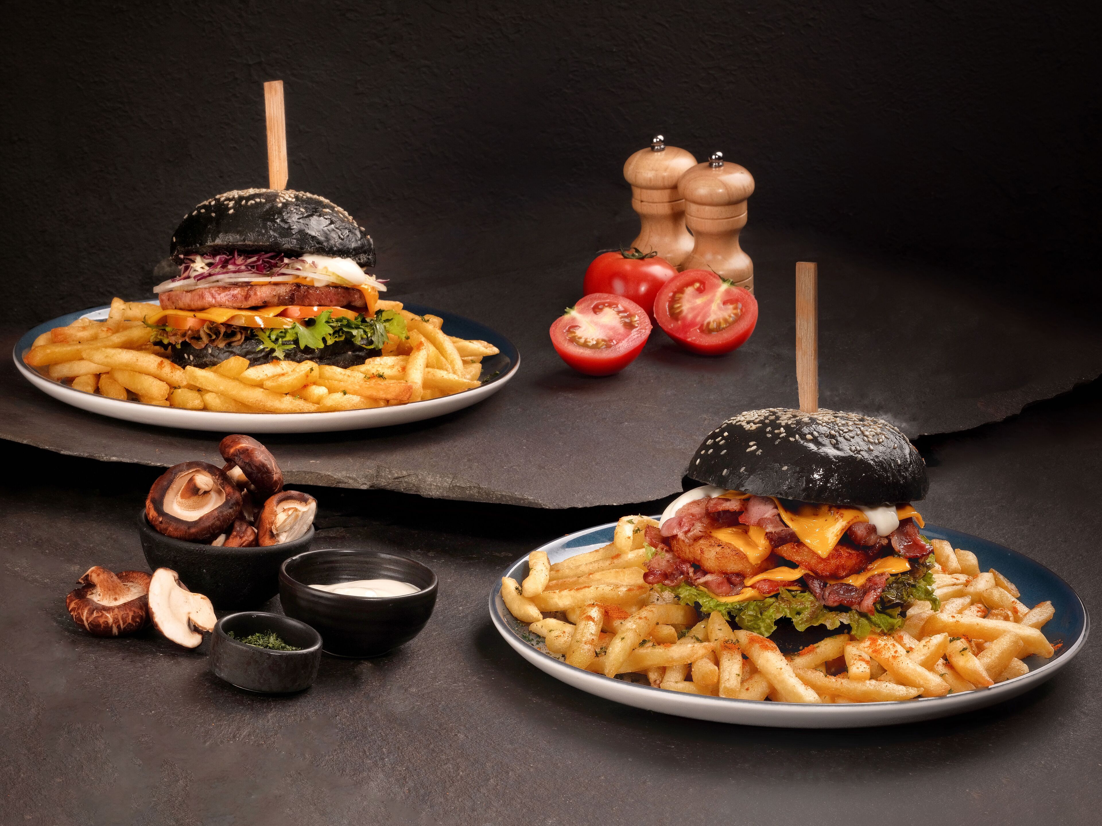 Dos hamburguesas con papas fritas en platos, un molinillo de pimienta y tomates.