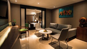 A modern hotel room with a sofa, chairs, a small round table with fruit, a wooden desk, and a painting on the wall.