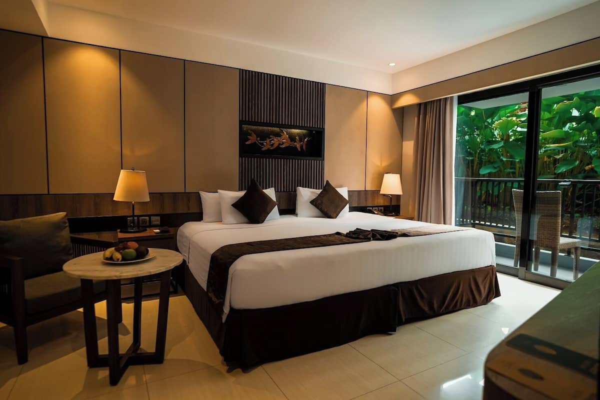 Ein modernes Hotelzimmer mit einem großen Bett, zwei Nachttischen mit Lampen, einem kleinen runden Tisch mit Obst und Blick auf Grün durch das Fenster.