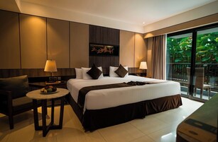 Habitación de hotel moderna con una cama grande, dos mesitas de noche con lámparas, una pequeña mesa redonda con fruta y vistas a zonas verdes por la ventana.