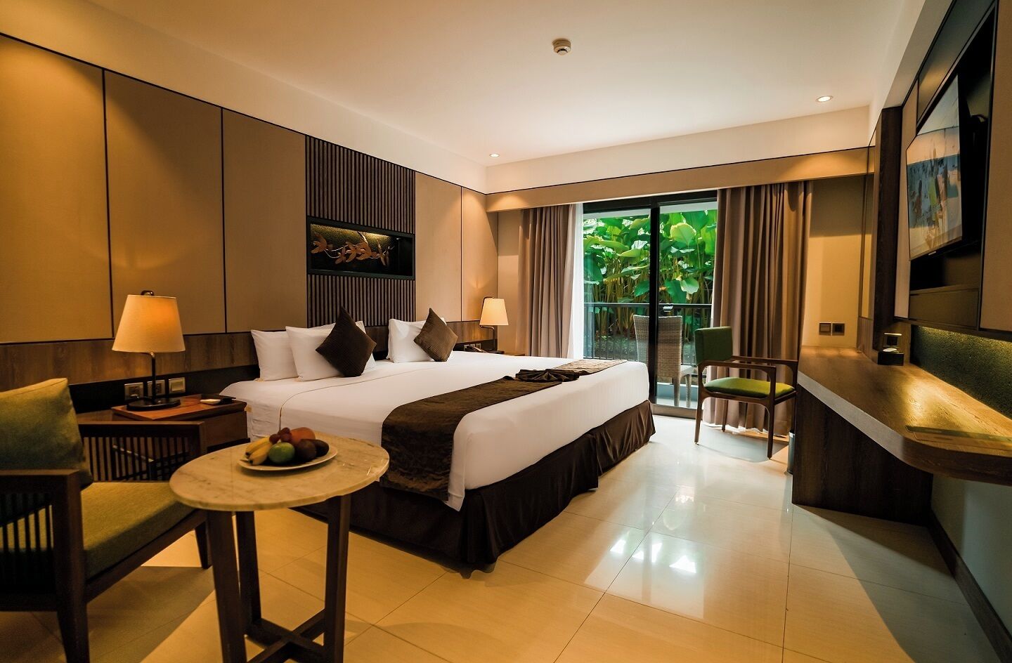 A modern hotel room with a large bed, a small dining table with fruit, a seating area with a chair, and a view of greenery through the window.