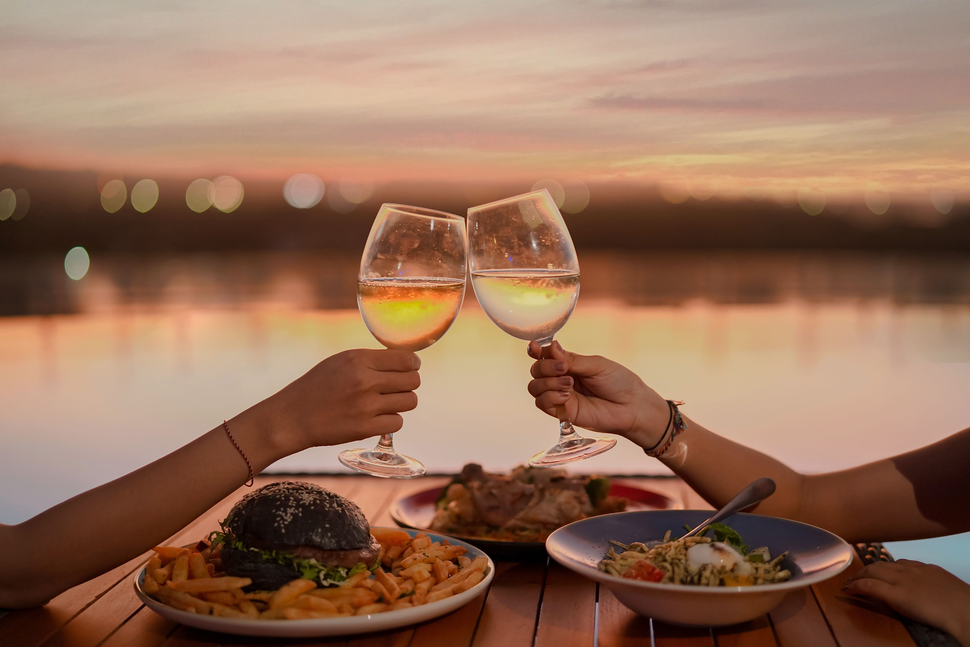 Mesa com comida e dois copos de vinho, com o pôr do sol ao fundo.