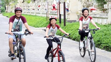 Three people riding bicycles on a paved path with greenery and a white fence in the background.