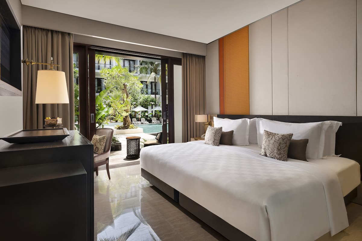 A modern hotel room with a large bed, a desk, and a view of the outdoor pool and greenery.